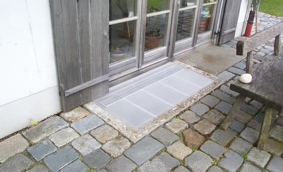 Transparente Lichtschachtabdeckung bündig eingelassen vor einer rustikalen Terrassentür mit Holzelementen.